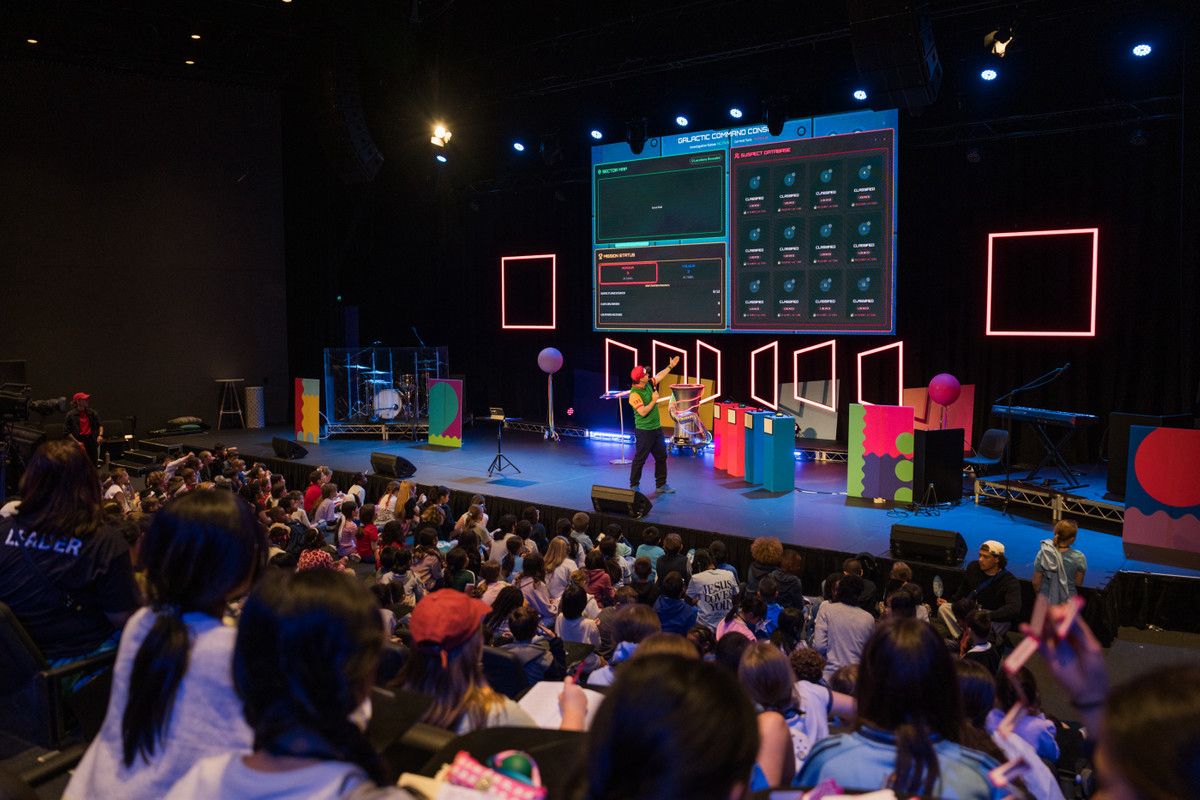 A church kids event seen from the back of the room: a colourful stage, screens, and a seated crowd facing forward.