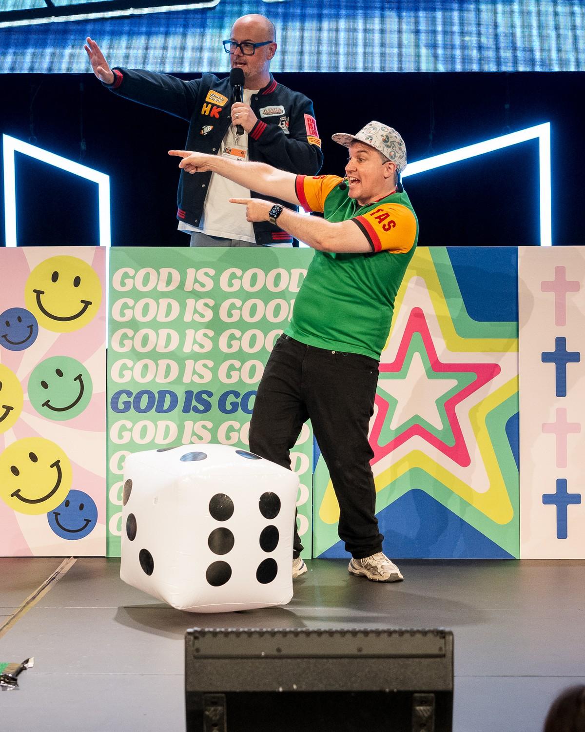 Presenters on a colourful church kids-event stage with a giant die and God is good backdrop.